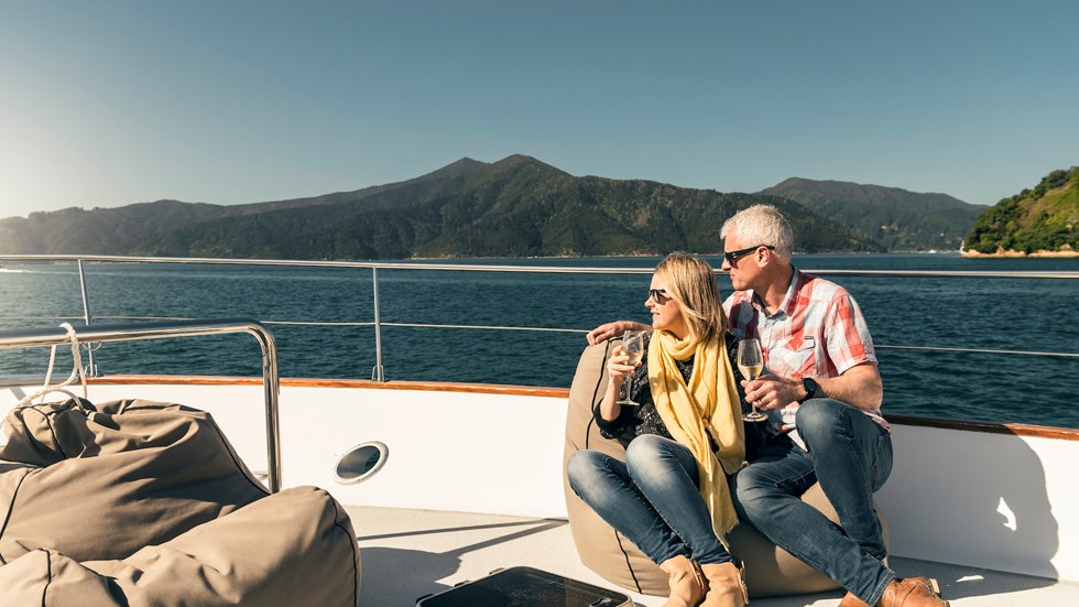Couple sit at the bow onboard a Marlborough Tour Company boat in the Marlborough Sounds at the top of New Zealand's South Island with glasses of wine looking at the view