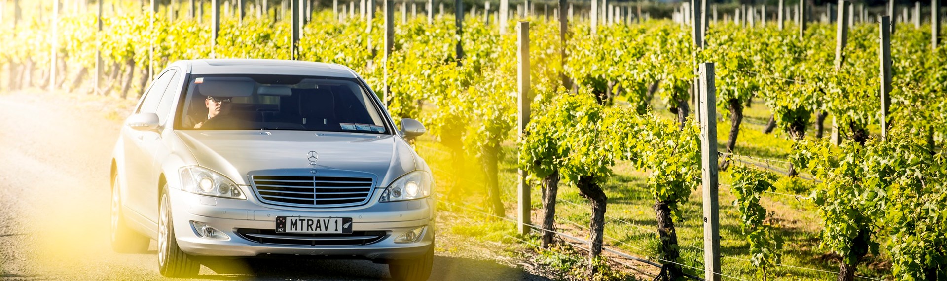 Silver car drives alongside a vineyard with hills in the background as part of a private wine tour in Marlborough near Blenheim, at the top of New Zealand's South Island