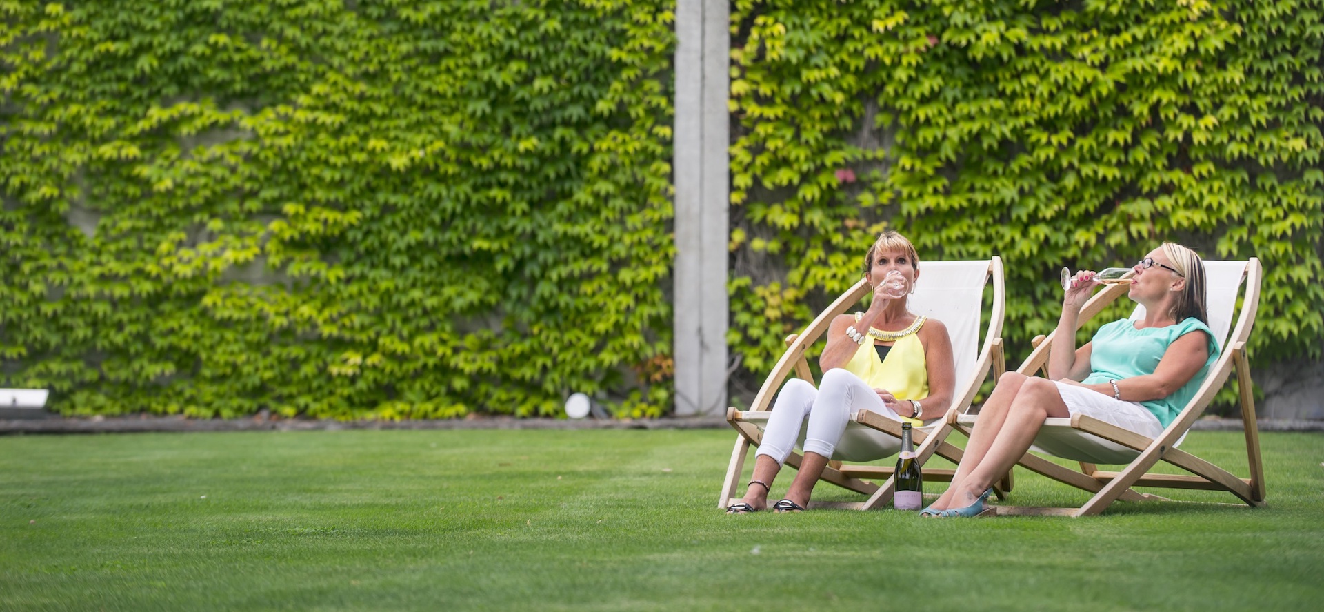 Two women on deck chairs on a lawn drinking wine at a winery cellar door in Marlborough, at the top of New Zealand's South Island