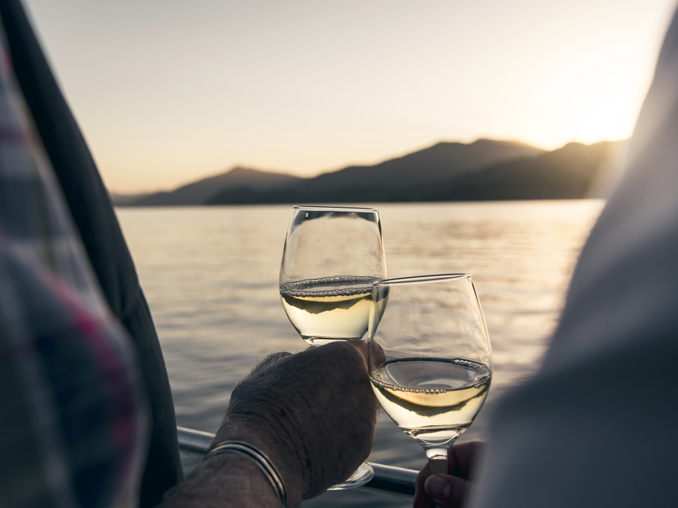 Close up of hands holding wine glasses with the Marlborough Sounds in the background at sunset - the top of New Zealand's South Island
