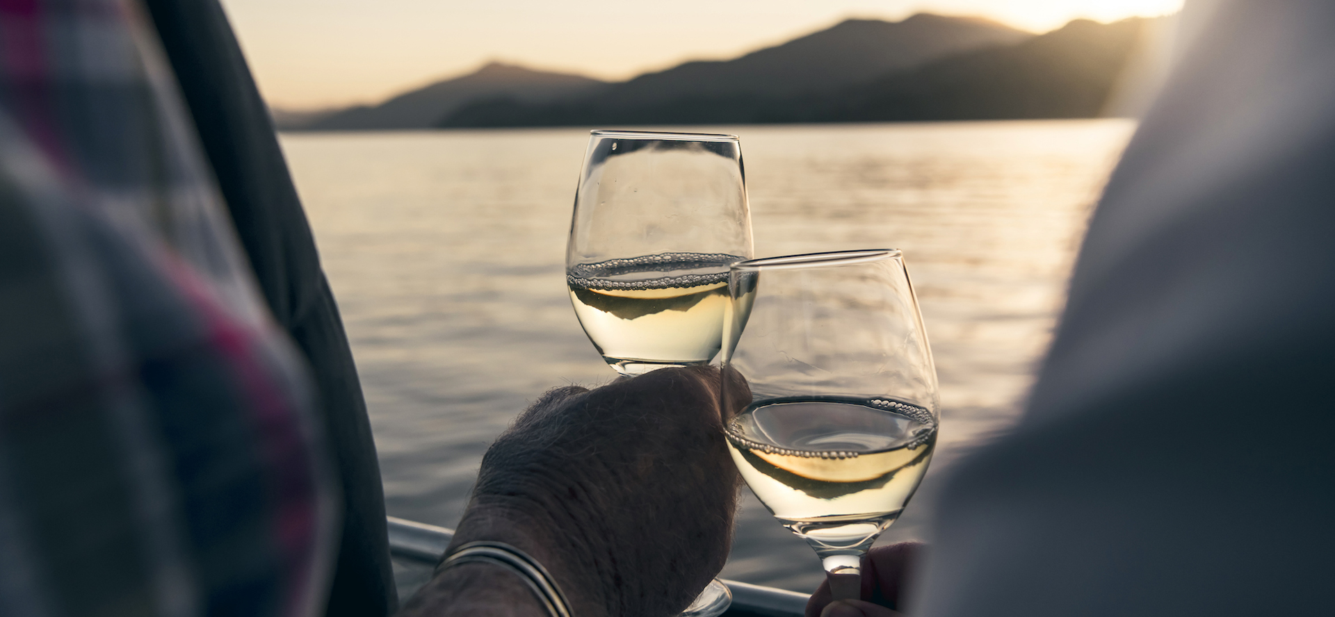 Close up of hands holding wine glasses with the Marlborough Sounds in the background at sunset - the top of New Zealand's South Island