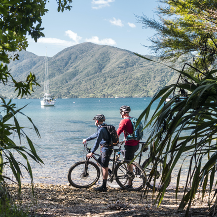 Two male cyclists take a rest beside the water with a yacht and Endeavour Inlet in the background.