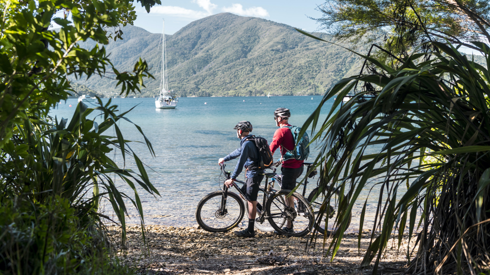 Two male cyclists take a rest beside the water with a yacht and Endeavour Inlet in the background.