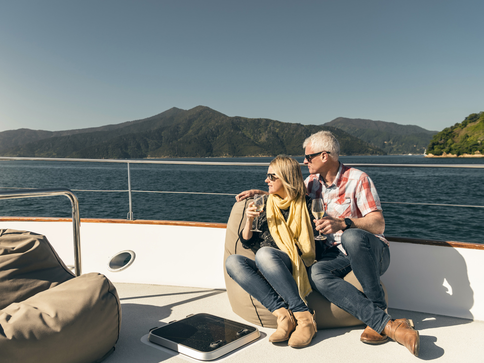 Couple sit at the bow onboard a Marlborough Tour Company boat in the Marlborough Sounds at the top of New Zealand's South Island with glasses of wine looking at the view