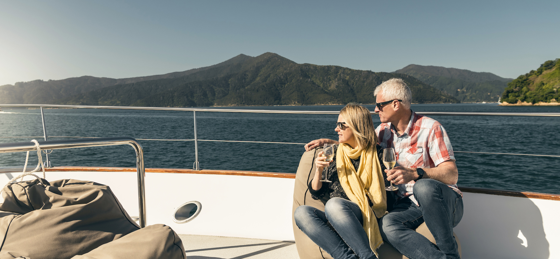 Couple sit at the bow onboard a Marlborough Tour Company boat in the Marlborough Sounds at the top of New Zealand's South Island with glasses of wine looking at the view