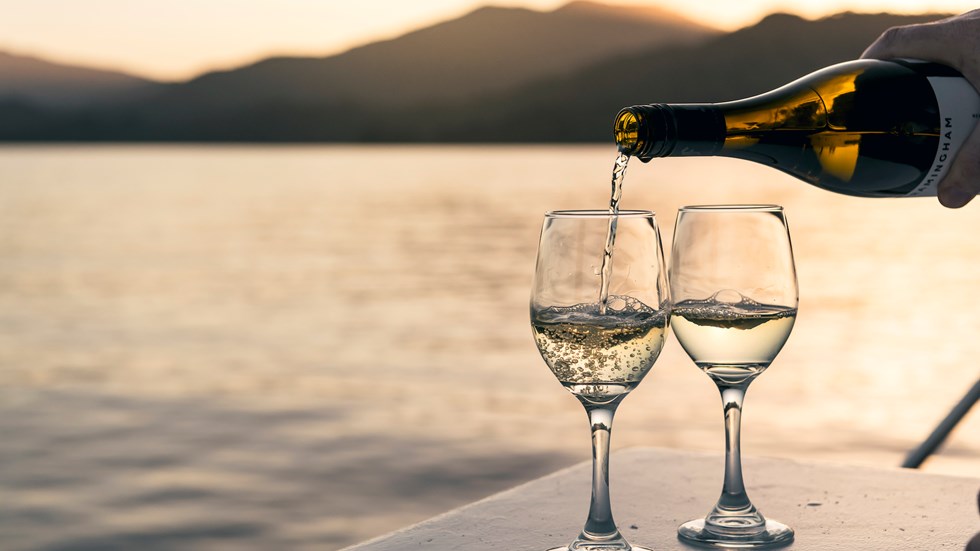 Wine bottle pouring white wine into two glasses at sunset onboard a boat in the Marlborough Sounds, at the top of New Zealand's South Island.