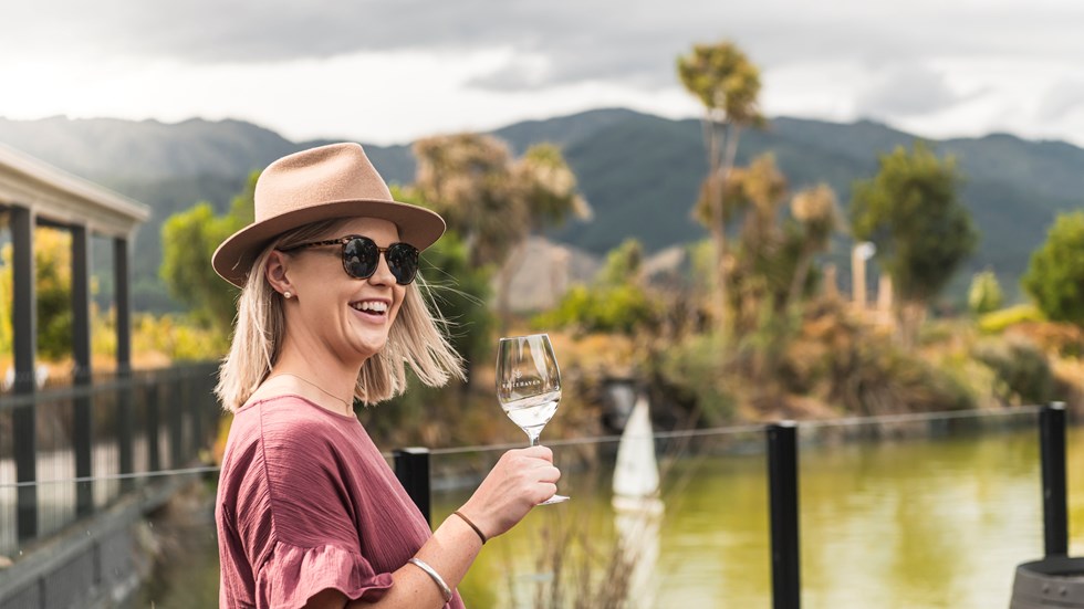 Woman with glass of wine outside in front of a pond with hill in background on a wine tour in Marlborough, at the top of New Zealand's South Island