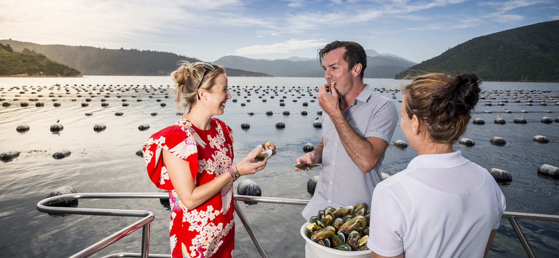 A couple laughs while tasting Greenshell mussels with Marlborough Tour Company staff member at bow of boat in the Marlborough Sounds, at the top of New Zealand's South Island