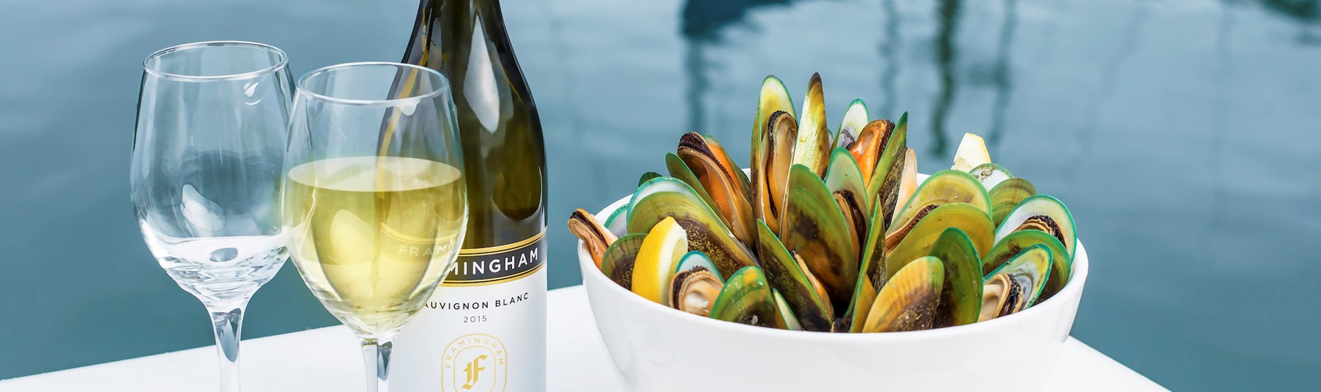 Two glasses of wine, a bottle of wine and bowl of freshly steamed mussels with the Marlborough Sounds in the background, at the top of New Zealand's South Island