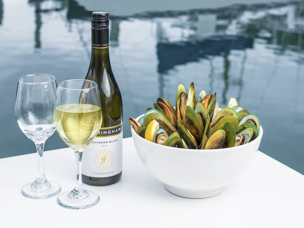 Two glasses of wine, a bottle of wine and bowl of freshly steamed mussels with the Marlborough Sounds in the background, at the top of New Zealand's South Island