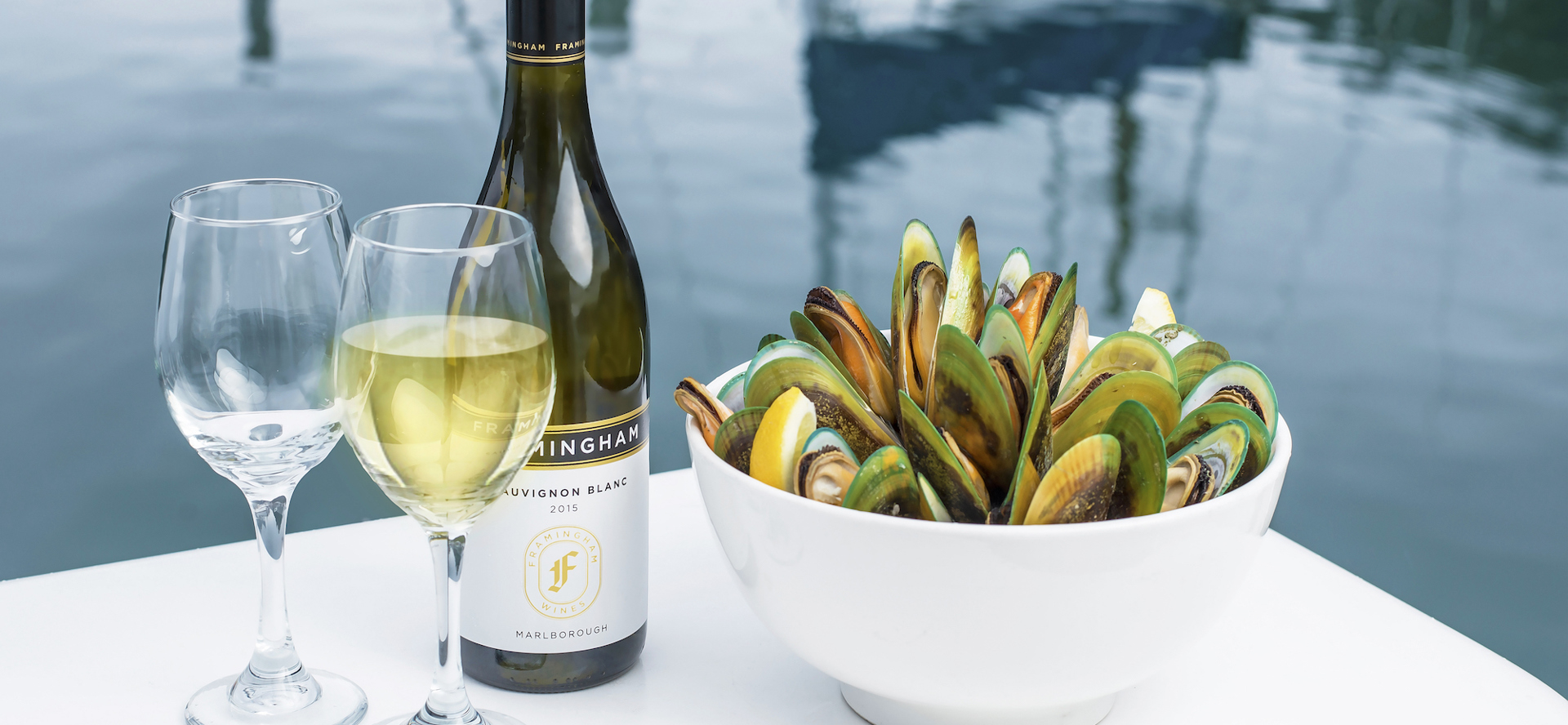 Two glasses of wine, a bottle of wine and bowl of freshly steamed mussels with the Marlborough Sounds in the background, at the top of New Zealand's South Island