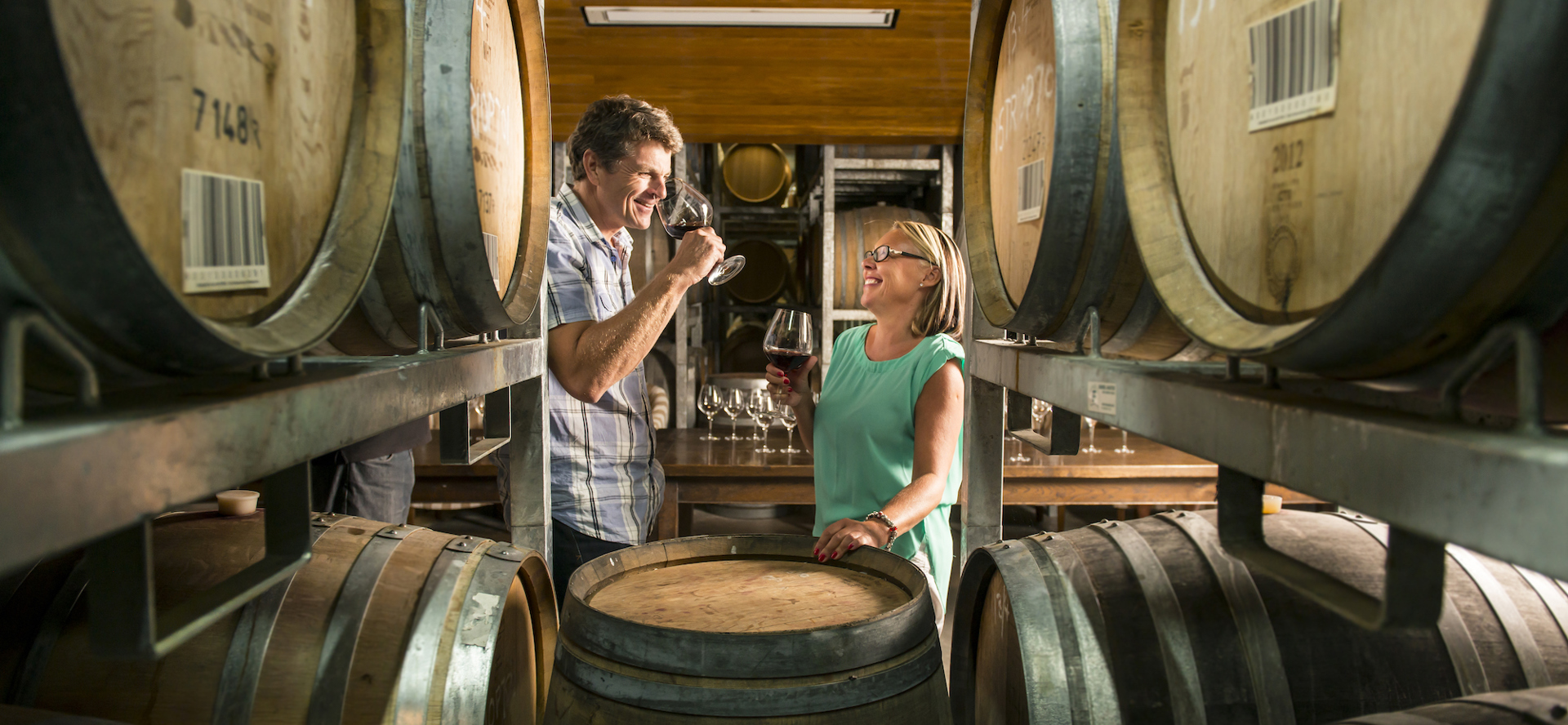 Couple drink red wine surrounded by wine barrels in a winery barrel hall on a wine tour in Marlborough near Blenheim, at the top of New Zealand's South Island