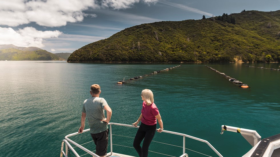 Couple on the bow of a boat looking at the Marlborough Sounds and mussel farm at the top of New Zealand's South Island