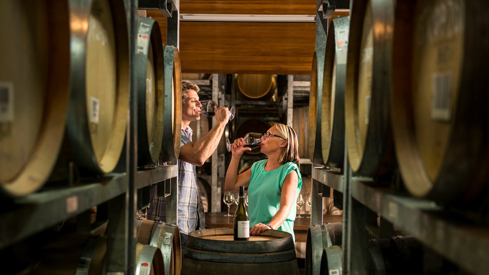 Couple doing a red wine tasting amongst wine barrels in a winery as part of a wine tour in Marlborough, as the top of New Zealand's South Island