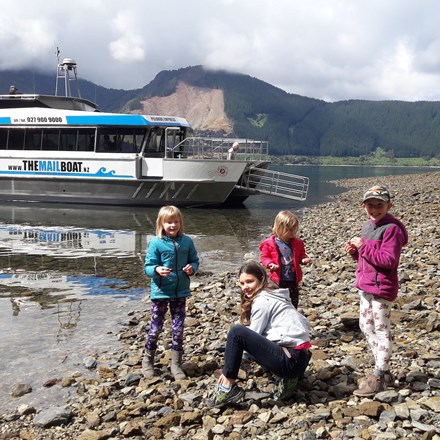 Four children on a rocky beach with Pelorus Mail Boat docked behind.